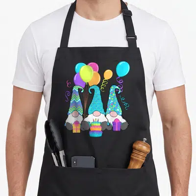 Gnome cake apron - Three gnomes with patterned hats holding cake and gifts under colorful balloons — shown on a black cotton canvas apron against a white background