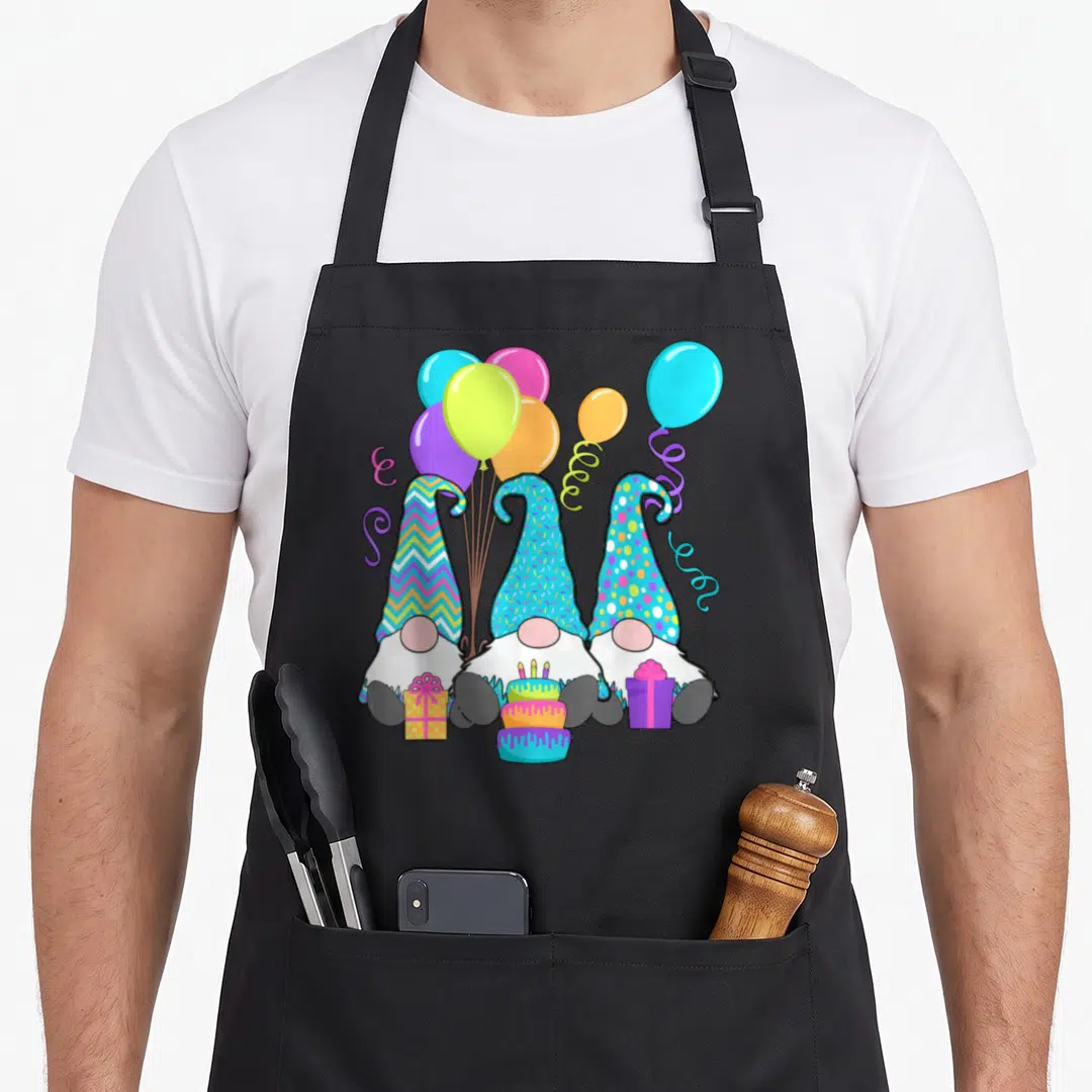 Gnome cake apron - Three gnomes with patterned hats holding cake and gifts under colorful balloons — shown on a black cotton canvas apron against a white background