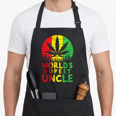 Dopest hemp lover apron - Grey cannabis leaf in red yellow green circle with forest base and stacked WORLD'S DOPEST — shown on a black cotton canvas apron against a white background