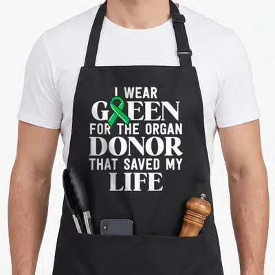 Transplant survivor organ apron - Vertical white text I WEAR GREEN FOR THE ORGAN DONOR THAT SAVED MY LIFE with green — shown on a black cotton canvas apron against a white background