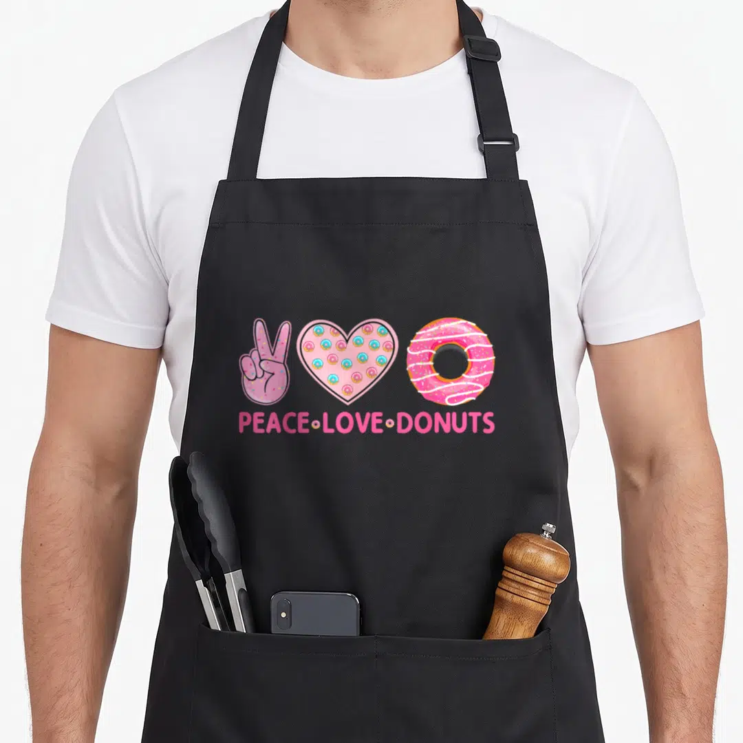 Themed donut apron - Cartoon peace sign, donut-filled heart, and glazed ring with pink text below — shown on a black cotton canvas apron against a white background