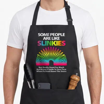 Slinky sarcastic apron - Rainbow slinky toy with white and gradient text saying some people are like slinkies — shown on a black cotton canvas apron against a white background