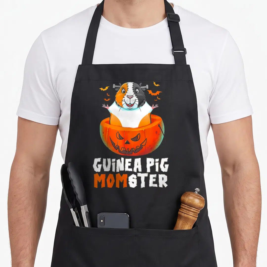 Guinea halloween apron - Cartoon guinea pig frankenstein in orange pumpkin with bats and stacked text — shown on a black cotton canvas apron against a white background