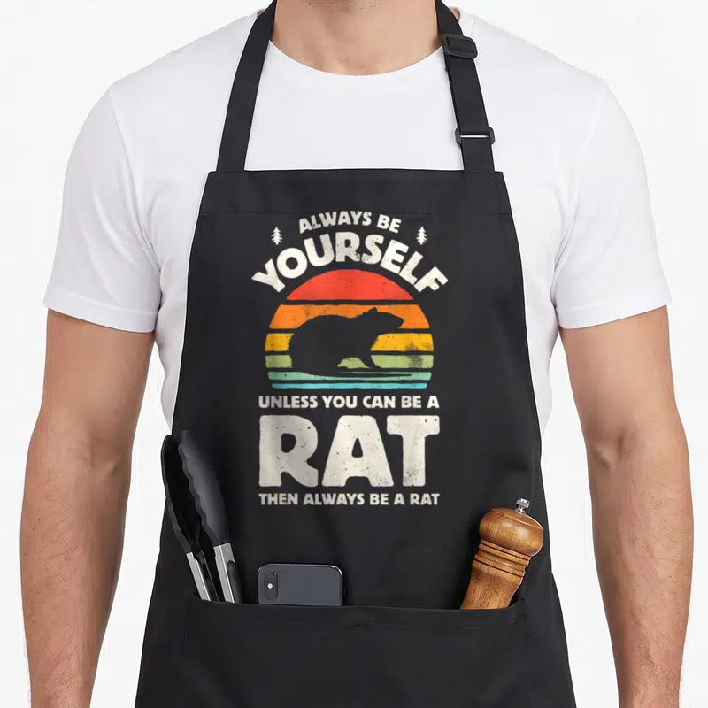 Alway funny rats apron - Rat silhouette against striped retro sun with sarcastic text about being a rodent — shown on a black cotton canvas apron against a white background