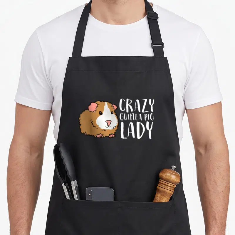 Funny guinea pet apron - Cartoon guinea pig in white brown pink next to three lines of text — shown on a black cotton canvas apron against a white background