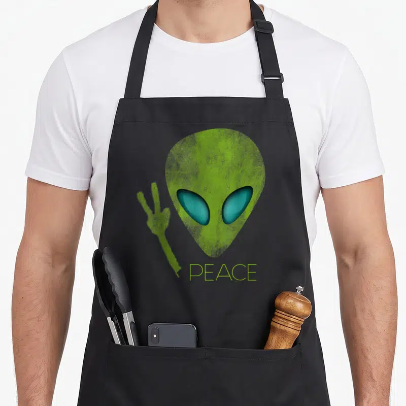 Alien birthday apron - Lime green alien head with teal eyes holding a peace sign above the word PEACE — shown on a black cotton canvas apron against a white background