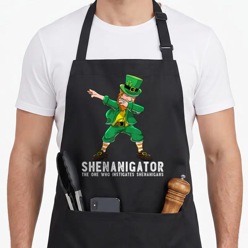 Shenanigator dabbing st patrick day aprons - Cartoon leprechaun dabbing with green hat and text reading Shenanigator — shown on a black cotton canvas apron against a white background