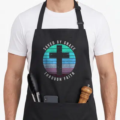 Through grace apron - Grey cross over teal blue purple striped circle with arched white text — shown on a black cotton canvas apron against a white background