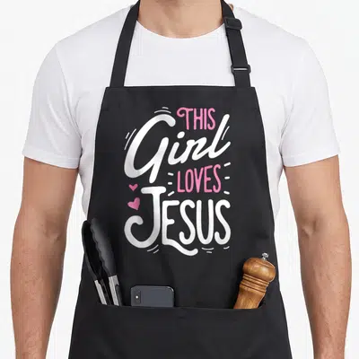 Jesus faith christian aprons - Stacked text reading This Girl Loves Jesus in white and pink with two small hearts — shown on a black cotton canvas apron against a white background