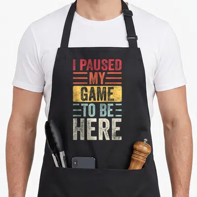 Paused gamer video game apron - Stacked I Paused My Game To Be Here text in red yellow blue white distressed font — shown on a black cotton canvas apron against a white background