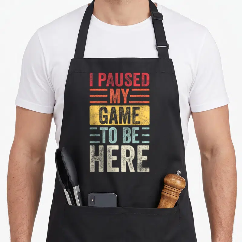 Paused gamer video game apron - Stacked I Paused My Game To Be Here text in red yellow blue white distressed font — shown on a black cotton canvas apron against a white background