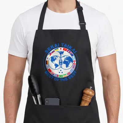 Cobra koki apron - Blue circular emblem with world map, karate kick, flags, and text sekai taikai best of the best — shown on a black cotton canvas apron against a white background