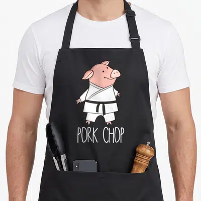 Pork animal apron - Pink cartoon pig in white karate uniform with black belt above PORK CHOP text — shown on a black cotton canvas apron against a white background