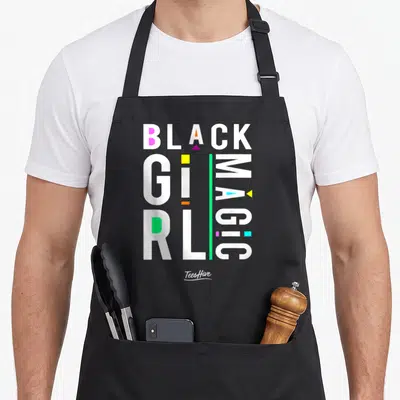 Afrocentric magic apron - Stacked BLACK GIRL MAGIC text with vertical MAGIC and scattered pink yellow orange green blue — shown on a black cotton canvas apron against a white background