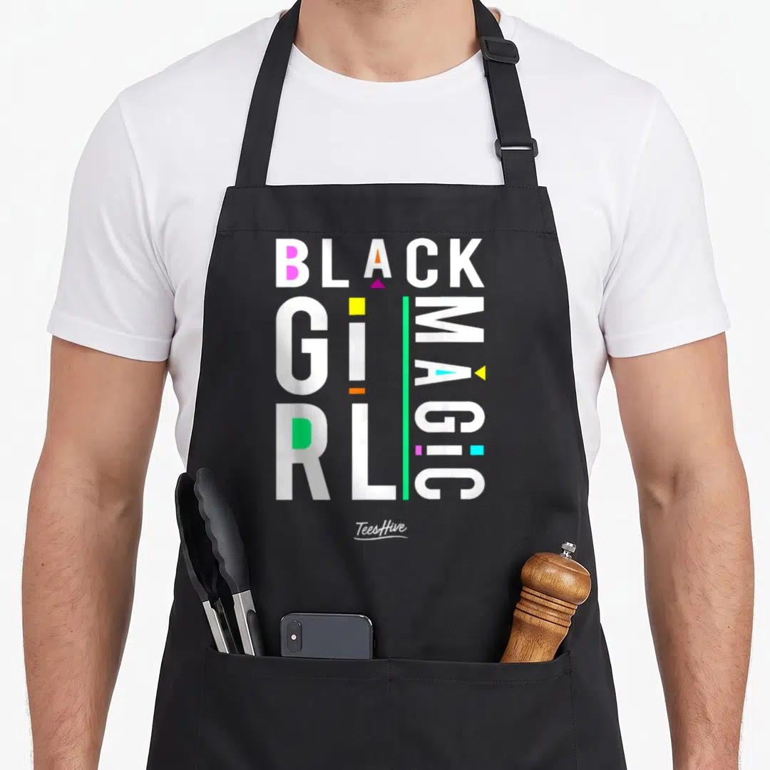 Afrocentric magic apron - Stacked BLACK GIRL MAGIC text with vertical MAGIC and scattered pink yellow orange green blue — shown on a black cotton canvas apron against a white background
