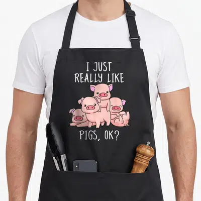 Swine pig lover apron - Four pink cartoon pigs stacked under text I JUST REALLY LIKE PIGS OK — shown on a black cotton canvas apron against a white background