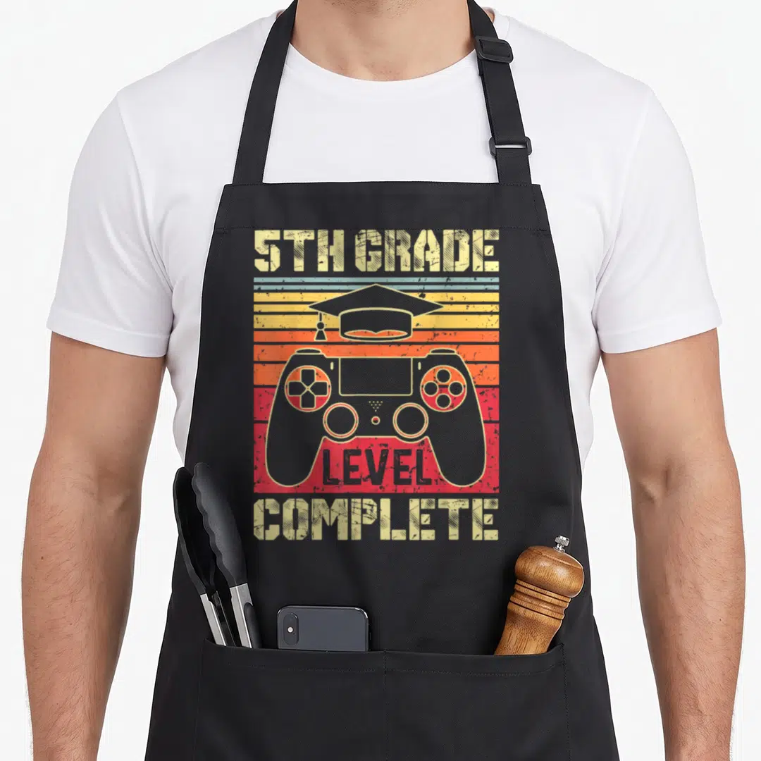 5th grad apron - Retro game controller with graduation cap and striped sunset background reading 5TH GRADE LEVEL — shown on a black cotton canvas apron against a white background