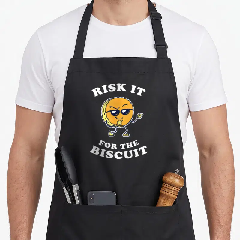 Risk chicken apron - Cartoon biscuit with sunglasses and Risk It For The Biscuit text in yellow white black — shown on a black cotton canvas apron against a white background