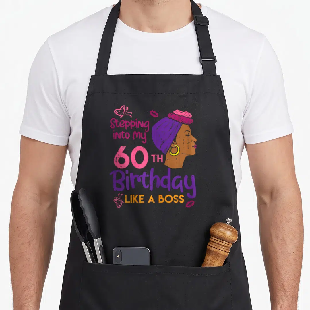 60th bbq boss apron - Cartoon woman profile with Stepping into my 60TH Birthday LIKE A BOSS text in purple and pink — shown on a black cotton canvas apron against a white background