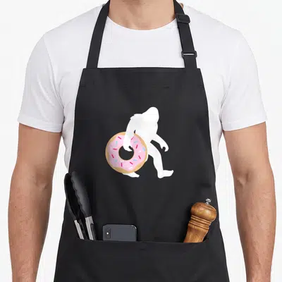 Bigfoot sprinkle apron - White Bigfoot silhouette holding a pink frosted donut with tan dough and sprinkles — shown on a black cotton canvas apron against a white background