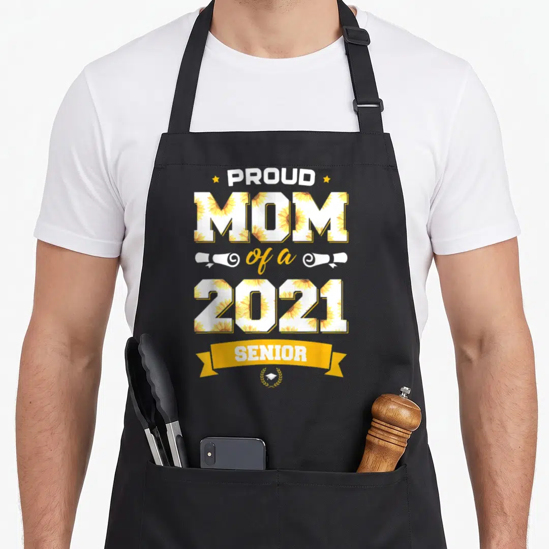Senior floral apron - Stacked PROUD MOM 2021 SENIOR text filled with yellow sunflowers and diplomas — shown on a black cotton canvas apron against a white background