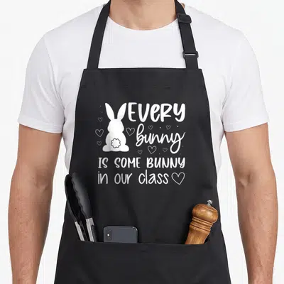 Our easter bunny aprons - White bunny silhouette next to stacked text reading EVERY bunny IS SOME BUNNY in our class — shown on a black cotton canvas apron against a white background