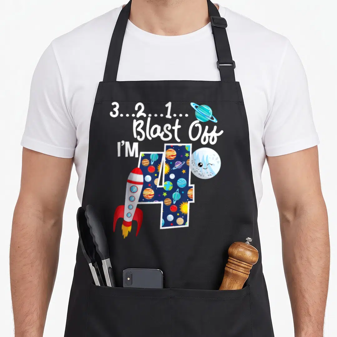 Blast birthday apron - Countdown text above galaxy-filled number 4 with rocket and winking planet — shown on a black cotton canvas apron against a white background