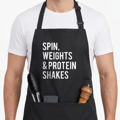 Weight spinning apron - White stacked text reading SPIN WEIGHTS & PROTEIN SHAKES on grey background — shown on a black cotton canvas apron against a white background