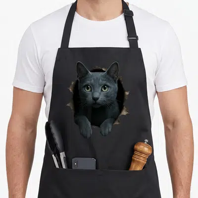 Adorable russian aprons - Grey cat face and paws emerging from a torn brown hole on a grey background — shown on a black cotton canvas apron against a white background