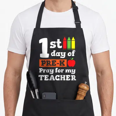 Pre teacher apron - Stacked white text reading 1st day of Pre-K Pray for my Teacher with three crayons and red apple — shown on a black cotton canvas apron against a white background