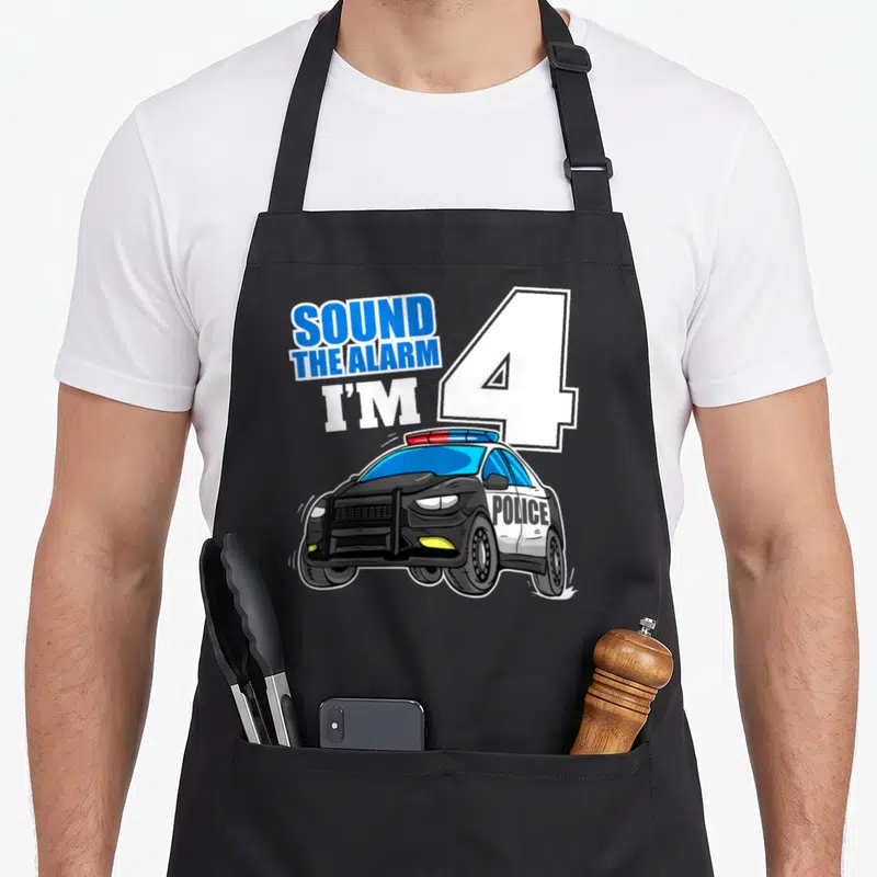 Police car apron - SOUND THE ALARM I'M 4 text with cartoon police car in blue and white — shown on a black cotton canvas apron against a white background