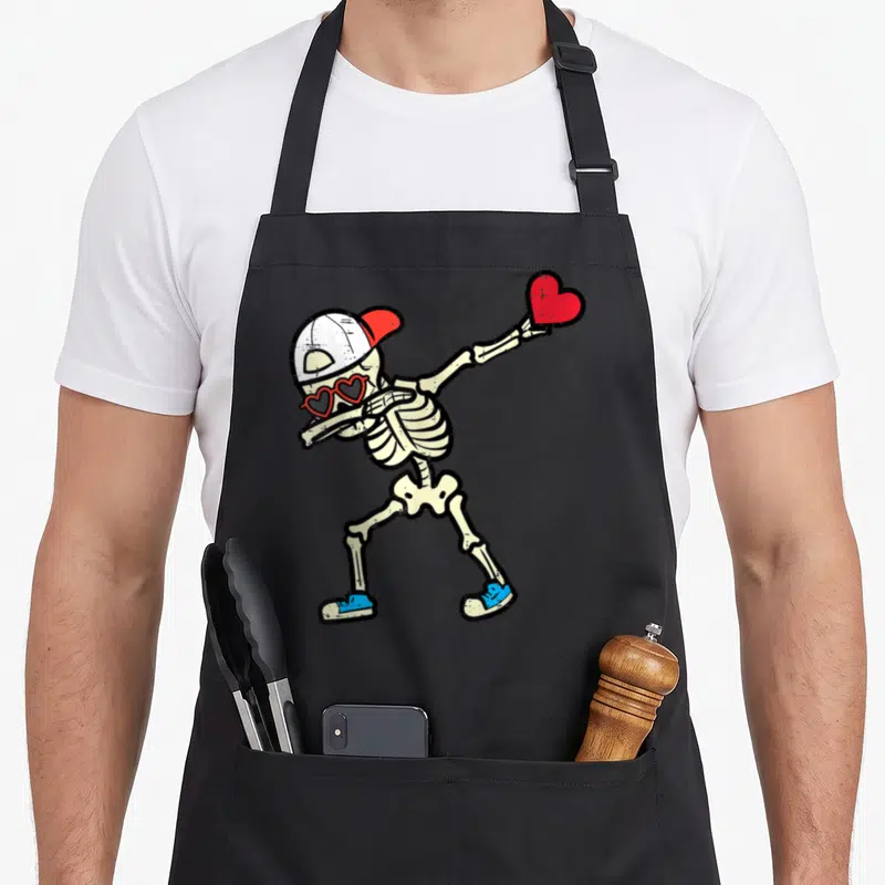 Skeleton valentine's day apron - Dabbing skeleton with heart sunglasses holding red heart, distressed print — shown on a black cotton canvas apron against a white background