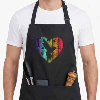 Distressed wooden apron - Black hummingbird silhouette inside rainbow wooden heart with paint splatters — shown on a black cotton canvas apron against a white background