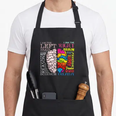 Brain parts of an apron - Split brain graphic with left reason words and right passion words in white pink yellow red — shown on a black cotton canvas apron against a white background