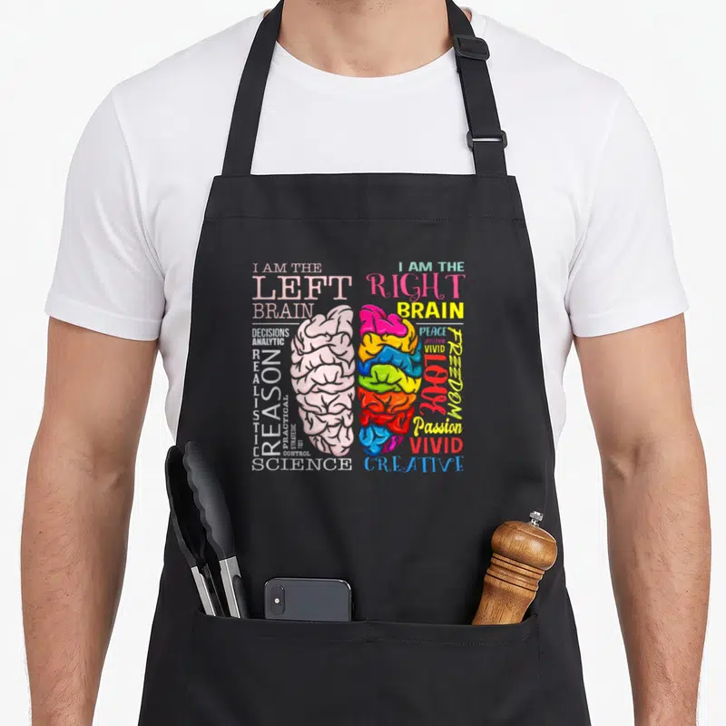 Brain parts of an apron - Split brain graphic with left reason words and right passion words in white pink yellow red — shown on a black cotton canvas apron against a white background