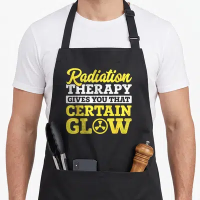 Beaten radiation aprons - Yellow and white stacked text reading Radiation Therapy Gives You That Certain Glow — shown on a black cotton canvas apron against a white background