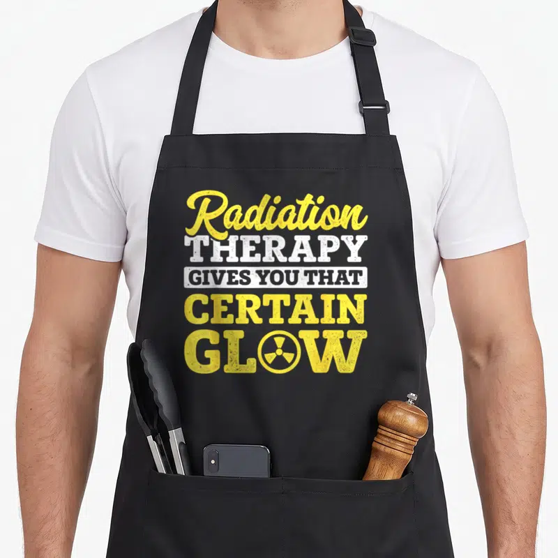 Beaten radiation aprons - Yellow and white stacked text reading Radiation Therapy Gives You That Certain Glow — shown on a black cotton canvas apron against a white background