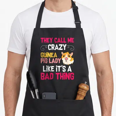 Call crazy guinea lady apron - Stacked colorful text THEY CALL ME CRAZY GUINEA PIG LADY with cartoon guinea pig — shown on a black cotton canvas apron against a white background