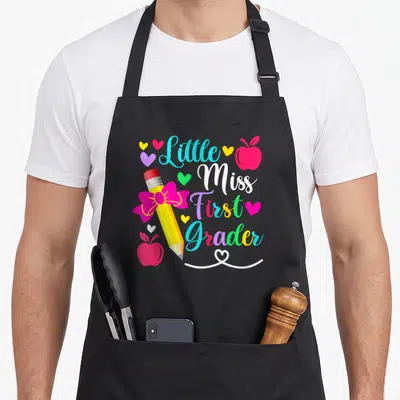 Grader miss grade apron - Little Miss First Grader text with pencil, apples, and hearts — shown on a black cotton canvas apron against a white background