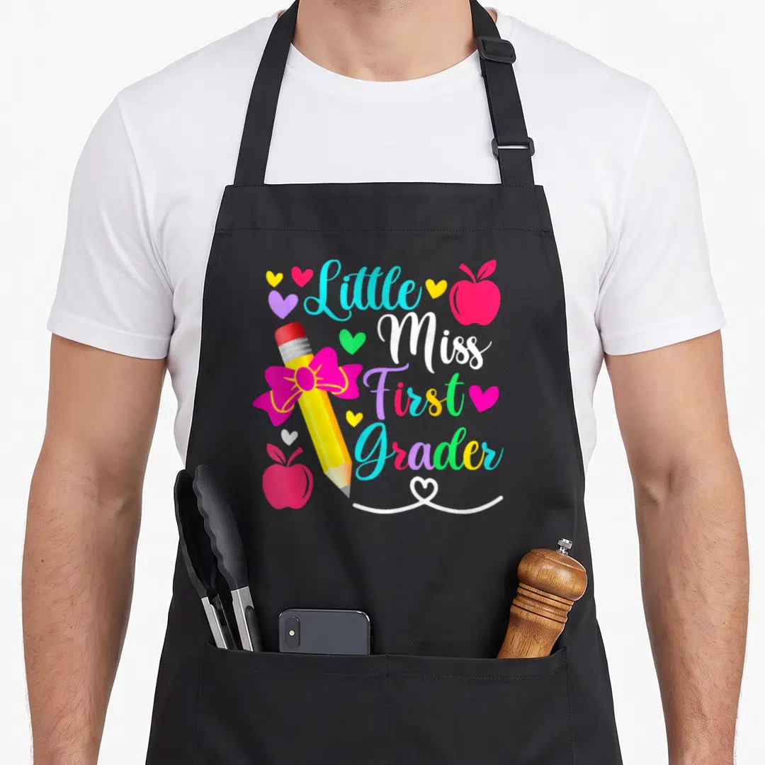 Grader miss grade apron - Little Miss First Grader text with pencil, apples, and hearts — shown on a black cotton canvas apron against a white background