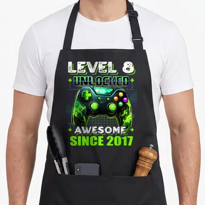 8th birthday level gamer video game apron - Game controller over blue grid with text LEVEL 8 UNLOCKED AWESOME SINCE 2017 — shown on a black cotton canvas apron against a white background