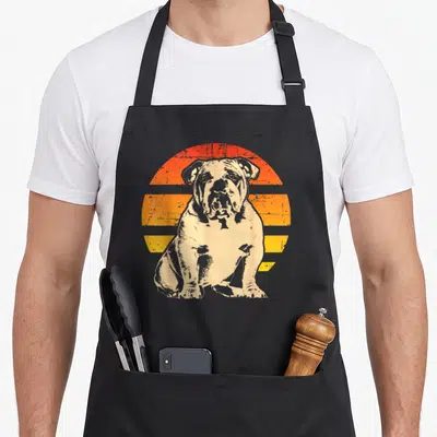 Funny bulldog english chef aprons - Distressed beige bulldog sitting in front of orange and yellow striped sun graphic — shown on a black cotton canvas apron against a white background