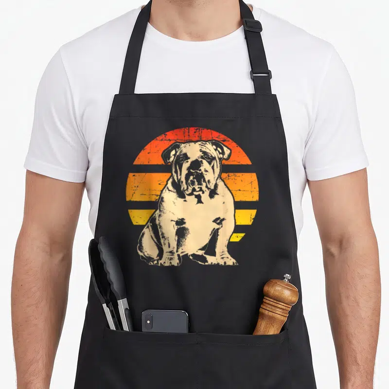 Funny bulldog english chef aprons - Distressed beige bulldog sitting in front of orange and yellow striped sun graphic — shown on a black cotton canvas apron against a white background