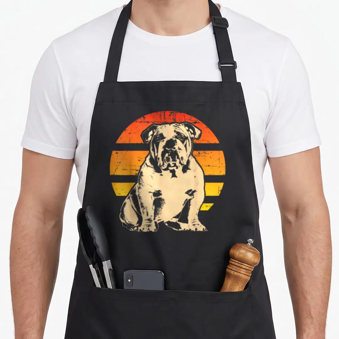 Funny bulldog english chef aprons - Distressed beige bulldog sitting in front of orange and yellow striped sun graphic — shown on a black cotton canvas apron against a white background