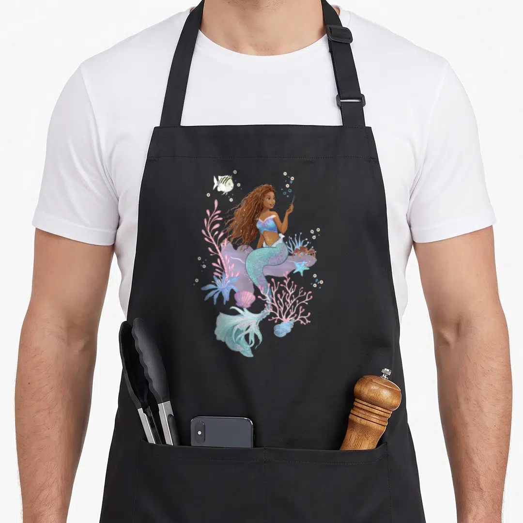 Flounder mermaid apron - Cartoon mermaid with teal tail, pink coral, blue fish, and brown crab illustration — shown on a black cotton canvas apron against a white background