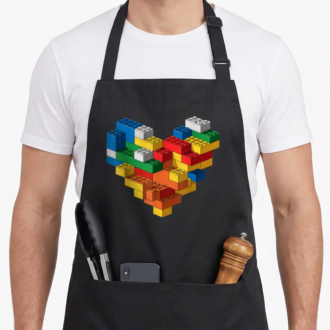 Brick builder building apron - Realistic heart shape built from blue, red, yellow, green, and white interlocking toy — shown on a black cotton canvas apron against a white background