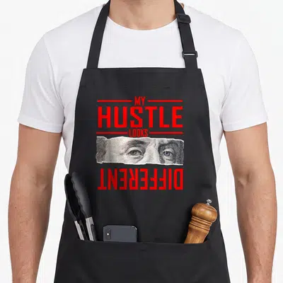 Different hustle apron - Red text reading MY HUSTLE LOOKS DIFFERENT above a rectangular cutout of realistic eyes — shown on a black cotton canvas apron against a white background