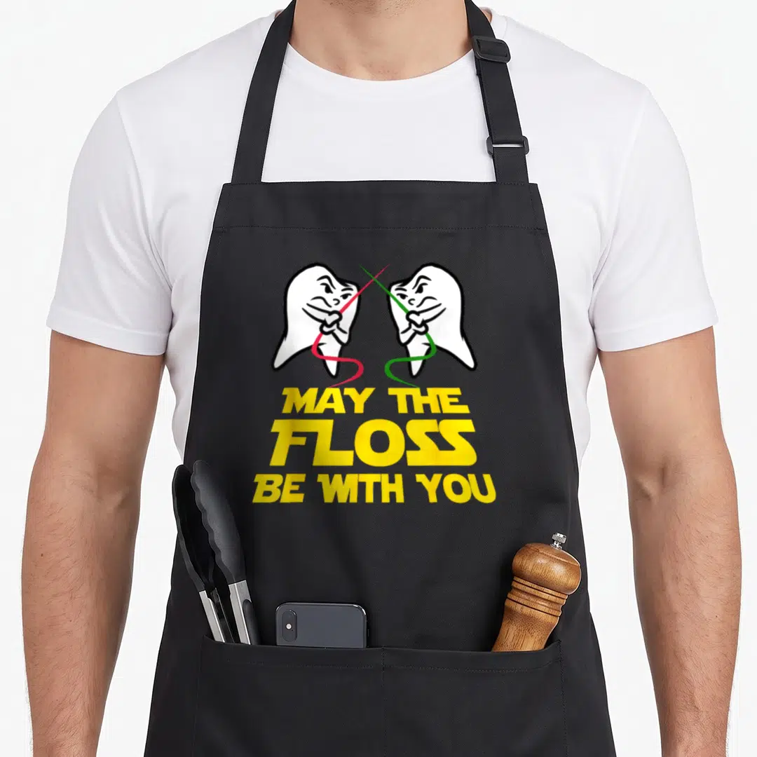 May tooth apron - Two cartoon teeth fencing with red and green floss above yellow May The Floss Be With You text — shown on a black cotton canvas apron against a white background