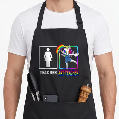 Dabbing teacher apron - Two frames showing a woman symbol labeled Teacher and a unicorn painting a rainbow labeled Art — shown on a black cotton canvas apron against a white background