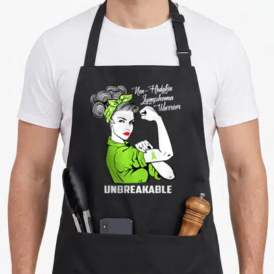 Non limes apron - Cartoon woman flexing arm with green ribbon and text Non-Hodgkin Lymphoma Warrior Unbreakable — shown on a black cotton canvas apron against a white background